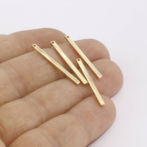 May include: A collection of gold-coloured rectangular metal bars, each with a small hole at one end. These thin bars, likely jewellery findings, are arranged against a white backdrop. The bars are approximately 2.5 cm in length.