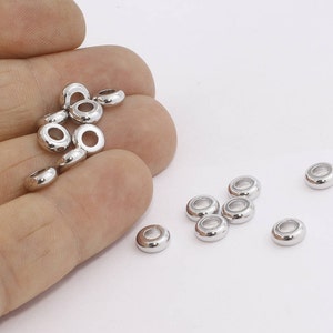 May include: A collection of shiny, silver-colored, donut-shaped beads. The beads are small, with a smooth, rounded surface and a hole in the center. They are scattered on a white background, some held in a hand.