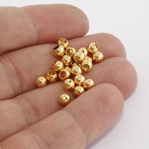 May include: A selection of small, shiny, gold beads. Each bead is spherical with a hole through the centre, designed for threading. The beads are shown in an open hand against a white background, suggesting use in jewellery making or crafts.