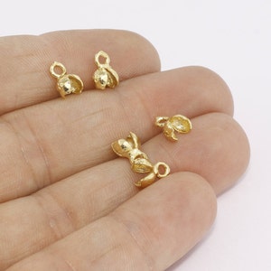 May include: Close-up of several small, gold-colored jewelry findings. The findings have a flower-like shape with a loop at the top for attaching to other jewelry components. The findings are on a light background.