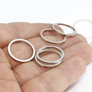 May include: A collection of silver-colored rings in various sizes and shapes. Some rings are round, while others are oval. The rings are held in a hand against a white background. The rings appear to be made of metal.