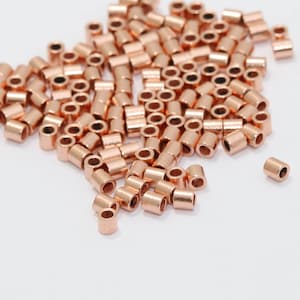 Może przedstawiać: Kolekcja małych, miedzianych metalowych koralików w kształcie tuby rozrzuconych na białej powierzchni. Cylindryczne koraliki mają otwór pośrodku, idealne do tworzenia biżuterii lub projektów rzemieślniczych.