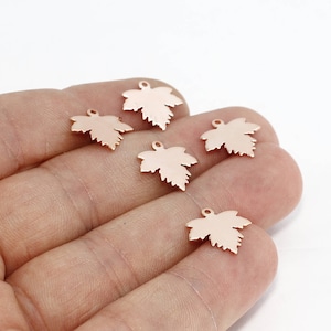 May include: Six small, rose gold-colored maple leaf charms. Each charm has a detailed leaf shape with a small loop at the top for attaching to jewelry or other crafts. The charms are displayed against a white background.