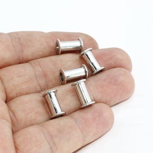 Op de afbeelding: Een hand die verschillende kleine, zilverkleurige metalen buiskralen vasthoudt. Elke kraal heeft een cilindrisch lichaam met een uitlopende rand aan beide uiteinden. De kralen worden tegen een witte achtergrond getoond.