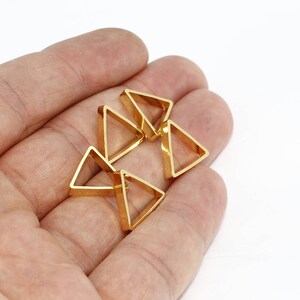 Puede incluir: Primer plano de seis pequeños dijes triangulares dorados. Los dijes se sostienen en una mano sobre un fondo blanco. Los dijes tienen un diseño geométrico y hueco, probablemente para la creación de joyas.
