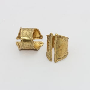 Könnte beinhalten: Zwei goldfarbene Ringe mit gehämmerten Texturen. Ein Ring ist intakt, der andere ist in zwei Teile geteilt. Die Ringe haben ein einzigartiges, geometrisches Design vor weißem Hintergrund.