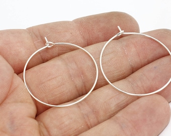 May include: Two silver hoop earrings. The earrings are thin and circular, with a simple, minimalist design. The hoops are held in a person's hand against a white background. The earrings are a classic jewelry item.