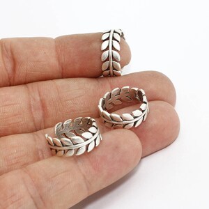 Puede incluir: Primer plano de varios anillos plateados con diseño de hojas. Los anillos están hechos de un metal brillante y presentan un patrón repetido de hojas alrededor de la banda. Los anillos se muestran sobre un fondo claro.