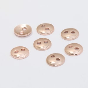 以下が含まれることがあります： 7つのローズゴールドのボタンのコレクション。各ボタンは丸く、2つの穴とわずかにドーム状の表面があります。ボタンは白い背景に配置され、金属的な輝きとシンプルなデザインが際立っています。縫製やクラフトプロジェクトに最適です。
