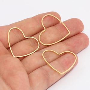 Puede incluir: Tres colgantes dorados con forma de corazón y estructura de alambre se muestran sobre un fondo blanco. Los colgantes son de varios tamaños y están sostenidos en la mano de una persona. Estos colgantes son adecuados para la fabricación de joyas.