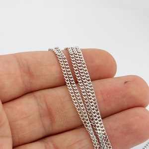May include: Close-up of several silver curb chains. The chains are displayed against a white background. The links are small and closely connected, creating a smooth, reflective surface. The chains are held in a hand.