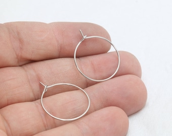 Könnte beinhalten: Zwei kleine, silberfarbene Creolen. Die Ohrringe sind kreisförmig und scheinen aus einem dünnen Metalldraht gefertigt zu sein. Die Ohrringe werden in einer Hand vor weißem Hintergrund gehalten.