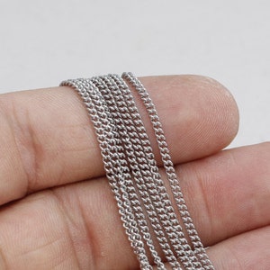 Könnte beinhalten: Nahaufnahme von mehreren Strängen einer silberfarbenen Panzerkette. Die Kette besteht aus kleinen, ineinandergreifenden Gliedern und wird in einer Hand vor weißem Hintergrund gehalten. Die Kette wird wahrscheinlich für die Schmuckherstellung verwendet.