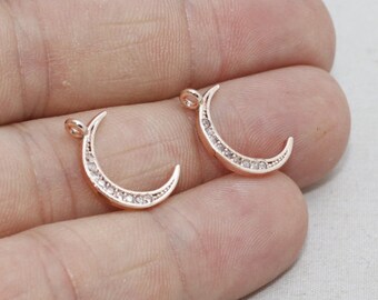 May include: Two rose gold crescent moon charms, each with a row of small, clear crystals along the curve. The charms have a small loop at the top for attaching to jewelry. The background is a light color.