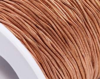 May include: A spool of copper-colored cord, tightly wound around a white plastic core. The cord has a metallic sheen, suggesting a decorative or craft material. The spool is set against a white background.