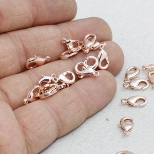 以下が含まれることがあります： ジュエリー作りに使用される、ローズゴールドカラーのロブスタークラスプが複数表示されています。 クラスプは小さく、湾曲したボディと、安全な閉鎖のためのスプリング式レバーを備えています。 明るい色の表面に散らばっています。