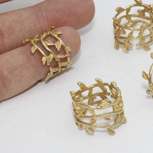 May include: Four gold-colored napkin rings with a leaf and vine design. One ring is on a finger. The rings are cylindrical with an open design, and the leaves are evenly spaced around the circumference.