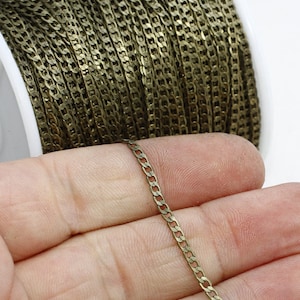 May include: A spool of antique brass-colored chain, with a small section held in a hand. The chain is a curb style, with a flat, rectangular link design. The chain is likely used for jewelry making or other crafts.