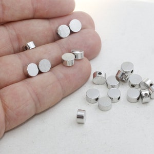 Könnte beinhalten: Eine Sammlung kleiner, glänzender, silberfarbener, runder Perlen. Jede Perle hat eine flache, kreisförmige Ober- und Unterseite mit einer zylindrischen Seite und einem kleinen Loch zum Auffädeln. Die Perlen sind verstreut, einige werden in einer Hand gehalten.