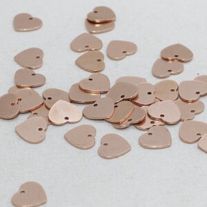 Puede incluir: Una colección de pequeños dijes en forma de corazón de color cobre dispersos sobre una superficie blanca. Cada dije tiene un pequeño agujero en la parte superior, lo que sugiere que están diseñados para la fabricación de joyas o manualidades. Los dijes tienen un acabado metálico liso.