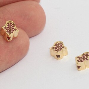 May include: Gold-colored Hamsa hand charms with red gemstone accents. The charms are small, with a detailed hand shape and a heart-shaped cutout. The red gemstones are set in a pattern on the palm of the hand.