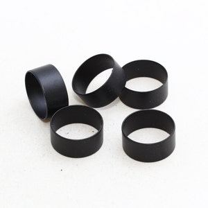 Puede incluir: Cinco servilleteros negros sobre un fondo blanco. Cada anillo es de forma cilíndrica, con un acabado mate. Están espaciados uniformemente, sugiriendo un conjunto para la mesa. Diámetro aproximado de 4 cm.