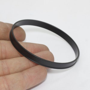 Pode incluir: Uma pulseira preta e circular com um acabamento liso e fosco. A pulseira tem um design de banda simples, adequado para uso diário. A imagem é definida contra um fundo branco liso.