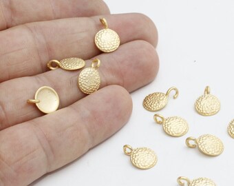 May include: A collection of gold-colored, textured metal pendants. The pendants are small, round, and oval-shaped with a hammered texture. Each pendant has a small loop at the top for attaching to a chain or other jewelry component. The pendants are scattered on a white surface.