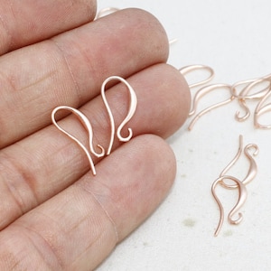 May include: Rose gold earring hooks, shown against a neutral background. The hooks have a simple, curved design with a looped end. Several hooks are visible, suggesting a bulk supply for jewellery making.