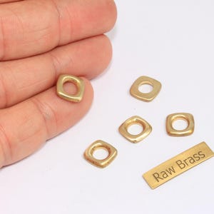 Op de afbeelding: Verschillende kleine, vierkante messing kralen met afgeronde hoeken en een centraal gat worden getoond. Een klein messing label met de woorden "Raw Brass" is ook aanwezig. De kralen hebben een warme goudkleur.