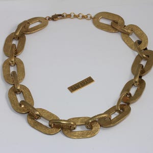Puede incluir: Un collar dorado con eslabones ovalados grandes y martillados. El collar está dispuesto en forma circular. También hay una pequeña etiqueta rectangular con el texto "RAW BRASS".