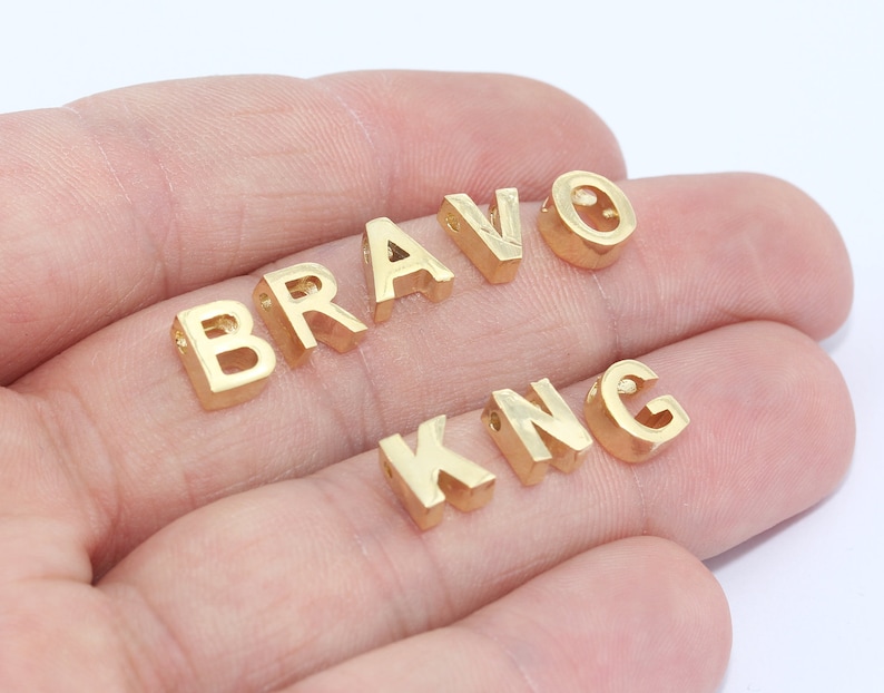 May include: Gold-colored metal letter beads spelling out "BRAVO" and "KNG". The letters are in a sans-serif font and have small holes for stringing. The beads are displayed against a white background.