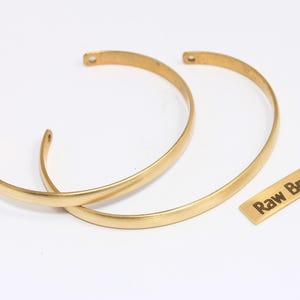 May include: Two gold-colored brass bangles and a small rectangular brass tag with the text "Raw Brass" are displayed on a white background. The bangles have a curved, open design.