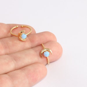 May include: Two gold-colored rings, one adjustable, featuring a crescent moon design. Each moon cradles a light blue opal-like stone. The rings are displayed on a light background.