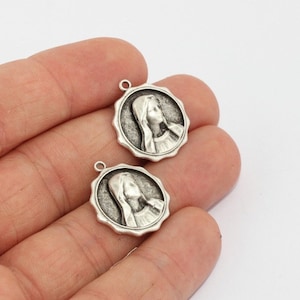 Puede incluir: Dos colgantes redondos plateados con un relieve detallado del perfil de una mujer. Cada colgante tiene un borde festoneado y un pequeño lazo en la parte superior para sujetarlo a una cadena u otro componente de joyería.