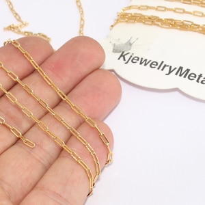 May include: Gold-colored chain with rectangular links, displayed on a white surface. The chain is held in a hand, with a tag that reads "KjewelryMetal". The chain is a bright, shiny gold color.
