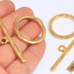 May include: Gold-plated toggle clasps and a ring clasp. The clasps are in a bright gold color. One of the toggle clasps is held in a hand. A small rectangular tag reads "24k Gold Plated".