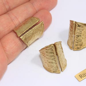 May include: Three raw brass rings with a hammered texture and a split design. The rings are gold-coloured and sit on a white surface. A small brass tag with the text "Raw Brass" is also present.