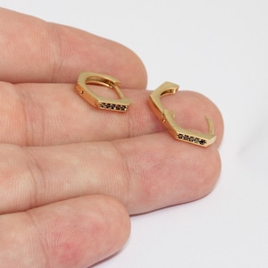 May include: A pair of gold-colored, geometric hoop earrings. Each earring features a series of small, dark-colored stones set along the bottom edge. The earrings are shown against a plain white background.