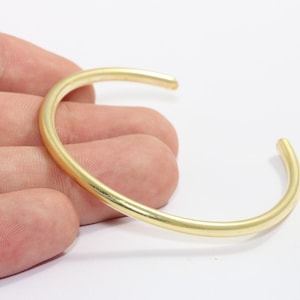 May include: A gold-colored, open cuff bracelet. The bracelet has a simple, rounded design and is held in a person's hand against a white background. The bracelet appears to be made of metal.