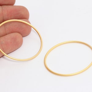 May include: Two gold-colored oval-shaped jewelry findings. The findings are thin, with a matte finish, and are displayed against a white background. These findings are likely used in jewelry making.