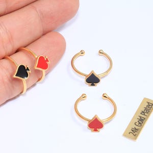 Könnte beinhalten: Vergoldete Ringe mit Spielkartenmotiven. Die Ringe zeigen schwarze Pik-, rote Herz- und rote Pik-Formen. Ein kleines rechteckiges Etikett lautet "24k Gold Plated".