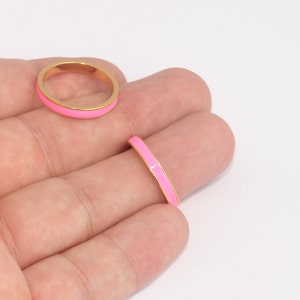 Puede incluir: Dos anillos dorados con una banda de esmalte rosa brillante. Un anillo está en un dedo y el otro está plano. Los anillos tienen un diseño simple y minimalista. El fondo es blanco.