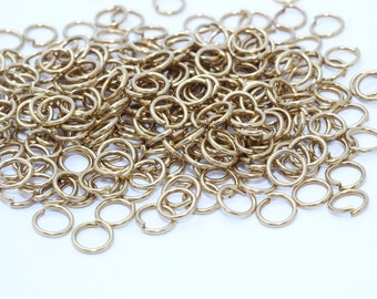 May include: A pile of gold-colored jump rings, each with a small opening. The rings are scattered on a white surface. These jewelry-making components are used for connecting various elements in jewelry designs.