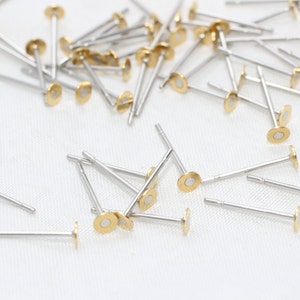 Op de afbeelding: Een verzameling kleine, zilverkleurige oorbelstiften met goudkleurige, platte, cirkelvormige toppen. De oorbellen zijn verspreid over een wit oppervlak, met veel in focus. De stiften zijn dun en hebben een eenvoudig, minimalistisch ontwerp.