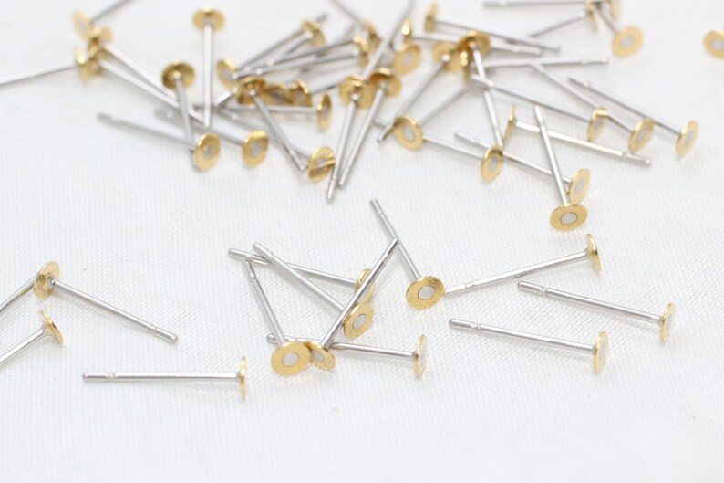Op de afbeelding: Een verzameling kleine, zilverkleurige oorbelstiften met goudkleurige, platte, cirkelvormige toppen. De oorbellen zijn verspreid over een wit oppervlak, met veel in focus. De stiften zijn dun en hebben een eenvoudig, minimalistisch ontwerp.