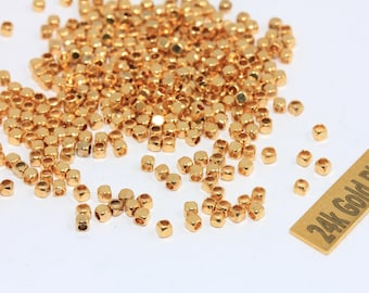 May include: A pile of small, gold-plated beads with a shiny finish. The beads are scattered on a white surface, with a small rectangular tag that reads "24k Gold Plated". These beads are ideal for jewelry making and crafting.