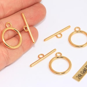 Könnte beinhalten: Vergoldete Schmuckteile, darunter Knebelverschlüsse und kreisförmige Ringe. Das Set enthält mehrere Knebelstangen und Ringe, ideal für die Schmuckherstellung. Ein kleines rechteckiges Stück trägt die Aufschrift "24k Gold Plated".