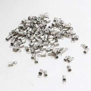 Puede incluir: Un conjunto de pequeños terminales de crimpado de color plateado, utilizados en la fabricación de joyas. Cada pieza tiene forma cilíndrica con un lazo en un extremo para la fijación. Los elementos están dispersos sobre una superficie blanca.