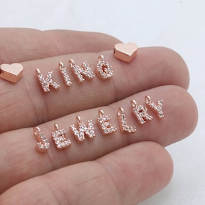 May include: Rose gold jewelry charms spelling out "KIND JEWELRY" with clear crystal accents. Includes two rose gold heart charms. These charms are perfect for creating personalized necklaces, bracelets, and other jewelry pieces.